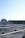A rooftop setting features a large satellite dish positioned prominently in the foreground. The roof is covered with a grid of white metal supports and scattered cables. In the background, there are various antennas and a structure with industrial elements, possibly a ventilation system. The sky is clear with a gradient of light blue, indicating a likely fair weather day.
