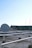 A rooftop setting features a large satellite dish positioned prominently in the foreground. The roof is covered with a grid of white metal supports and scattered cables. In the background, there are various antennas and a structure with industrial elements, possibly a ventilation system. The sky is clear with a gradient of light blue, indicating a likely fair weather day.