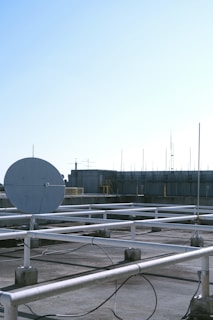 A rooftop setting features a large satellite dish positioned prominently in the foreground. The roof is covered with a grid of white metal supports and scattered cables. In the background, there are various antennas and a structure with industrial elements, possibly a ventilation system. The sky is clear with a gradient of light blue, indicating a likely fair weather day.