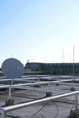 A rooftop setting features a large satellite dish positioned prominently in the foreground. The roof is covered with a grid of white metal supports and scattered cables. In the background, there are various antennas and a structure with industrial elements, possibly a ventilation system. The sky is clear with a gradient of light blue, indicating a likely fair weather day.