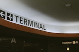 An indoor setting featuring a large sign indicating a terminal in an airport, accompanied by an airplane icon. An escalator sign with a directional arrow is visible nearby. The area is well-lit with muted colors.