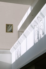 Sunlight creates intricate window frame shadows on a minimalist interior wall. A small framed picture hangs to the left, adding a subtle touch to the modern space. The architecture features clean lines and muted colors, enhancing the play of light and shadow.