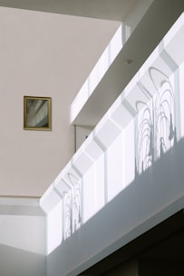 Sunlight creates intricate window frame shadows on a minimalist interior wall. A small framed picture hangs to the left, adding a subtle touch to the modern space. The architecture features clean lines and muted colors, enhancing the play of light and shadow.