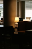A dimly lit room features a table lamp casting a warm glow on a nearby surface, surrounded by shadows. The setting appears to be a cozy indoor space, possibly a living room or lounge. Soft light filtering through window blinds in the background adds to the ambiance.