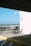 A lone wheelchair is positioned against a white wall with an attached camera, in an open parking area. Sunlight casts sharp shadows, and in the background, a clear view of an airport runway is visible along with parked cars.