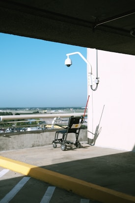 A lone wheelchair is positioned against a white wall with an attached camera, in an open parking area. Sunlight casts sharp shadows, and in the background, a clear view of an airport runway is visible along with parked cars.