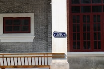A brick facade of a building features a white window frame with a smaller red window on the left and a large red-framed window on the right. A street sign labeled 'Changxing Road' in both English and Chinese is mounted on a white pillar. The building's wall consists of gray bricks and there is a wooden railing at the bottom.