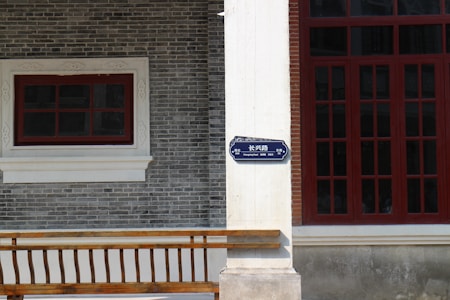 A brick facade of a building features a white window frame with a smaller red window on the left and a large red-framed window on the right. A street sign labeled 'Changxing Road' in both English and Chinese is mounted on a white pillar. The building's wall consists of gray bricks and there is a wooden railing at the bottom.