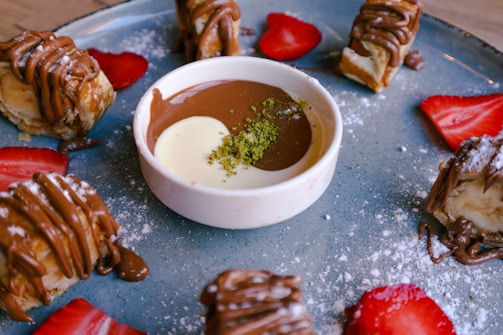 A dessert plate featuring a small bowl of two-tone sauce with a sprinkle of green garnish in the center, surrounded by banana slices wrapped in pastry and drizzled with chocolate, along with sliced strawberries.