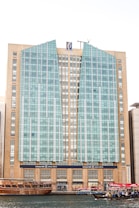 A tall, modern building with a distinctive facade composed of glass panels and beige stone sections. The architecture features vertical and angular elements, with a logo at the top center. In the foreground, traditional wooden boats and smaller vessels, some with UAE flags, float on a body of water, suggesting a mix of modern and traditional influences.