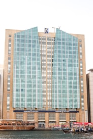 A tall, modern building with a distinctive facade composed of glass panels and beige stone sections. The architecture features vertical and angular elements, with a logo at the top center. In the foreground, traditional wooden boats and smaller vessels, some with UAE flags, float on a body of water, suggesting a mix of modern and traditional influences.