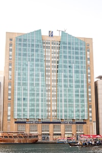 A tall, modern building with a distinctive facade composed of glass panels and beige stone sections. The architecture features vertical and angular elements, with a logo at the top center. In the foreground, traditional wooden boats and smaller vessels, some with UAE flags, float on a body of water, suggesting a mix of modern and traditional influences.