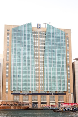 A tall, modern building with a distinctive facade composed of glass panels and beige stone sections. The architecture features vertical and angular elements, with a logo at the top center. In the foreground, traditional wooden boats and smaller vessels, some with UAE flags, float on a body of water, suggesting a mix of modern and traditional influences.