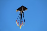 A kite with a unique design soars in the sky. The kite resembles a black square with prominent eye-like patterns. Below it, multiple streamers dangle downward, colored in yellow, red, and blue, against a clear blue sky.