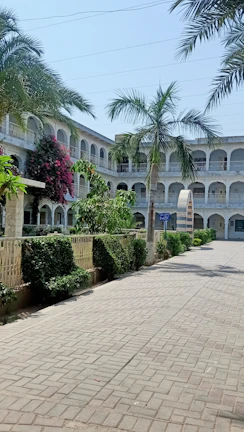 A courtyard surrounded by a two-story building with arches and balconies, lined with palm trees and various green plants. The pathway is paved with tiles and there is a vibrant pink flowering bush against one of the walls.