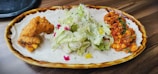 A plate of food featuring crispy fried chicken, a fresh green salad garnished with flowers and herbs, and a portion of spicy-looking orange and red dish, possibly a stir-fried or sauced mix.