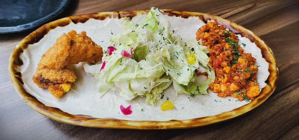 A plate of food featuring crispy fried chicken, a fresh green salad garnished with flowers and herbs, and a portion of spicy-looking orange and red dish, possibly a stir-fried or sauced mix.