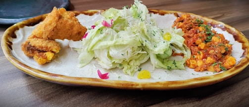 A plated dish featuring crispy fried chicken pieces on the left, with a pile of fresh, shredded lettuce and edible flowers in the center. On the right, there is a portion of a vibrant tomato and corn vegetable mixture. The dish sits on a glazed ceramic plate with a rustic brown edge.