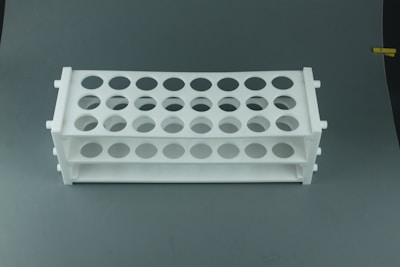 A white plastic test tube rack with multiple circular holes arranged in rows, set against a plain gray background.