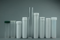 A collection of various cylindrical containers displayed in a row. The containers vary in size and include test tubes and storage tubes. They have different types of closures, such as screw caps, and are predominantly transparent or opaque white. The minimalist arrangement and neutral background create a clinical and organized appearance.