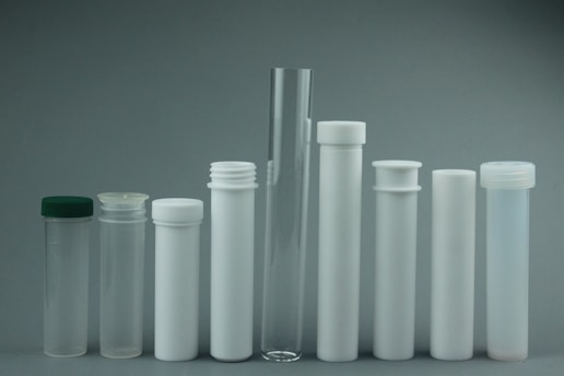 A collection of various cylindrical containers displayed in a row. The containers vary in size and include test tubes and storage tubes. They have different types of closures, such as screw caps, and are predominantly transparent or opaque white. The minimalist arrangement and neutral background create a clinical and organized appearance.