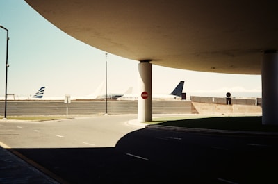 Several airplanes are parked at an airport, with their tails visible against the sky. The foreground includes a road with a 'no entry' sign and is partly in shadow due to an overhead structure. There are lamp posts and other airport infrastructure in the background.