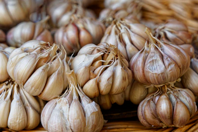 Fresh garlic bulbs with papery skins resting on a woven basket, surrounded by green leaves.