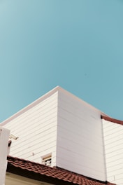 A minimalist architectural scene featuring the corner of a white modern building with clean lines is set against a bright blue sky. The building has a reddish-brown tiled roof, offering a contrast to the white walls and the sky. A small window is visible, partially open, on one of the walls.
