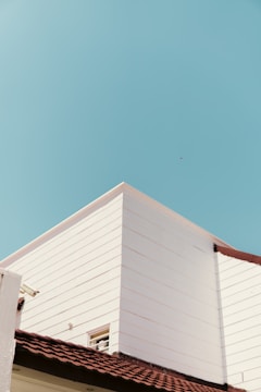 A minimalist architectural scene featuring the corner of a white modern building with clean lines is set against a bright blue sky. The building has a reddish-brown tiled roof, offering a contrast to the white walls and the sky. A small window is visible, partially open, on one of the walls.