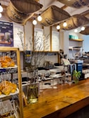 A cozy cafe interior featuring a wooden counter adorned with a vase of tall branches and a display of freshly baked croissants. Overhead, burlap coffee sacks are suspended alongside warm, glowing light bulbs. Behind the counter are various coffee-making tools and menu boards against a tiled wall.