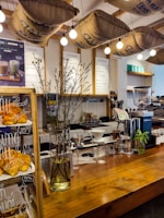A cozy cafe interior featuring a wooden counter adorned with a vase of tall branches and a display of freshly baked croissants. Overhead, burlap coffee sacks are suspended alongside warm, glowing light bulbs. Behind the counter are various coffee-making tools and menu boards against a tiled wall.