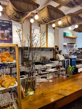 A cozy cafe interior featuring a wooden counter adorned with a vase of tall branches and a display of freshly baked croissants. Overhead, burlap coffee sacks are suspended alongside warm, glowing light bulbs. Behind the counter are various coffee-making tools and menu boards against a tiled wall.