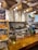 A cozy cafe interior featuring a wooden counter adorned with a vase of tall branches and a display of freshly baked croissants. Overhead, burlap coffee sacks are suspended alongside warm, glowing light bulbs. Behind the counter are various coffee-making tools and menu boards against a tiled wall.