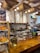 A cozy cafe interior featuring a wooden counter adorned with a vase of tall branches and a display of freshly baked croissants. Overhead, burlap coffee sacks are suspended alongside warm, glowing light bulbs. Behind the counter are various coffee-making tools and menu boards against a tiled wall.