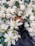 An arrangement of white roses, lilies, and various greenery is interspersed with small white flowers and larger round blooms. A black mesh-like material is present among the flowers, creating a contrast against the light colors.