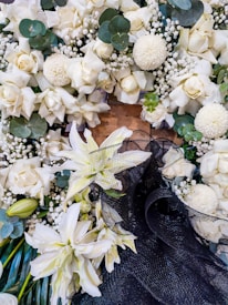 An arrangement of white roses, lilies, and various greenery is interspersed with small white flowers and larger round blooms. A black mesh-like material is present among the flowers, creating a contrast against the light colors.