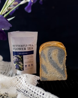 A package of butterfly pea flower tea is placed next to a loaf of marble bread, showcasing a swirl of blue and beige hues. The background is dark, and there are vibrant purple flowers in the upper corners. A white lace cloth is partially visible at the bottom.