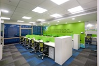 A modern office workspace with green and white color accents. The room features several workstations with green dividers and black swivel chairs. The walls are decorated with motivational phrases, and there is a green accent wall. The office has a grid-patterned carpet in shades of blue and dark grey, and large windows allowing natural light in.