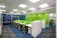 A modern office workspace with green and white color accents. The room features several workstations with green dividers and black swivel chairs. The walls are decorated with motivational phrases, and there is a green accent wall. The office has a grid-patterned carpet in shades of blue and dark grey, and large windows allowing natural light in.