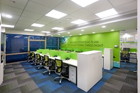 A modern office workspace with green and white color accents. The room features several workstations with green dividers and black swivel chairs. The walls are decorated with motivational phrases, and there is a green accent wall. The office has a grid-patterned carpet in shades of blue and dark grey, and large windows allowing natural light in.