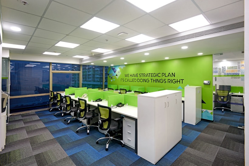 A modern office workspace with green and white color accents. The room features several workstations with green dividers and black swivel chairs. The walls are decorated with motivational phrases, and there is a green accent wall. The office has a grid-patterned carpet in shades of blue and dark grey, and large windows allowing natural light in.