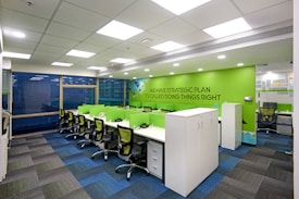 A modern office workspace with green and white color accents. The room features several workstations with green dividers and black swivel chairs. The walls are decorated with motivational phrases, and there is a green accent wall. The office has a grid-patterned carpet in shades of blue and dark grey, and large windows allowing natural light in.