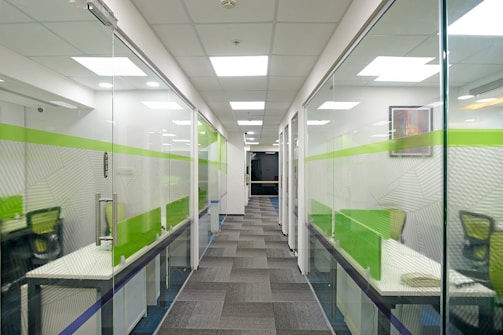 A modern office hallway with glass-walled cubicles on both sides, featuring bright overhead lighting. The cubicles have green and white design elements, with individual desks and chairs visible inside.