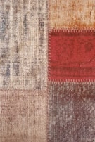 Close-up view of a textured fabric or carpet with a patchwork design consisting of four squares. The squares have different patterns and colors including beige, red, and brown. The red square has visible stitching along its borders.
