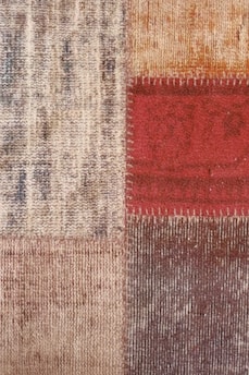 Close-up view of a textured fabric or carpet with a patchwork design consisting of four squares. The squares have different patterns and colors including beige, red, and brown. The red square has visible stitching along its borders.