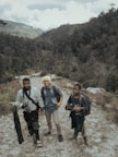 Three people with backpacks stand on a rugged path within a mountainous and forested landscape. The individuals appear to be prepared for an outdoor adventure or trek, carrying equipment such as a camera and tripod.