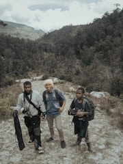 Three people with backpacks stand on a rugged path within a mountainous and forested landscape. The individuals appear to be prepared for an outdoor adventure or trek, carrying equipment such as a camera and tripod.