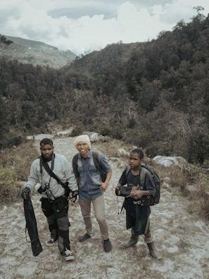 Three people with backpacks stand on a rugged path within a mountainous and forested landscape. The individuals appear to be prepared for an outdoor adventure or trek, carrying equipment such as a camera and tripod.