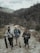 Three people with backpacks stand on a rugged path within a mountainous and forested landscape. The individuals appear to be prepared for an outdoor adventure or trek, carrying equipment such as a camera and tripod.