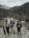 Three people with backpacks stand on a rugged path within a mountainous and forested landscape. The individuals appear to be prepared for an outdoor adventure or trek, carrying equipment such as a camera and tripod.